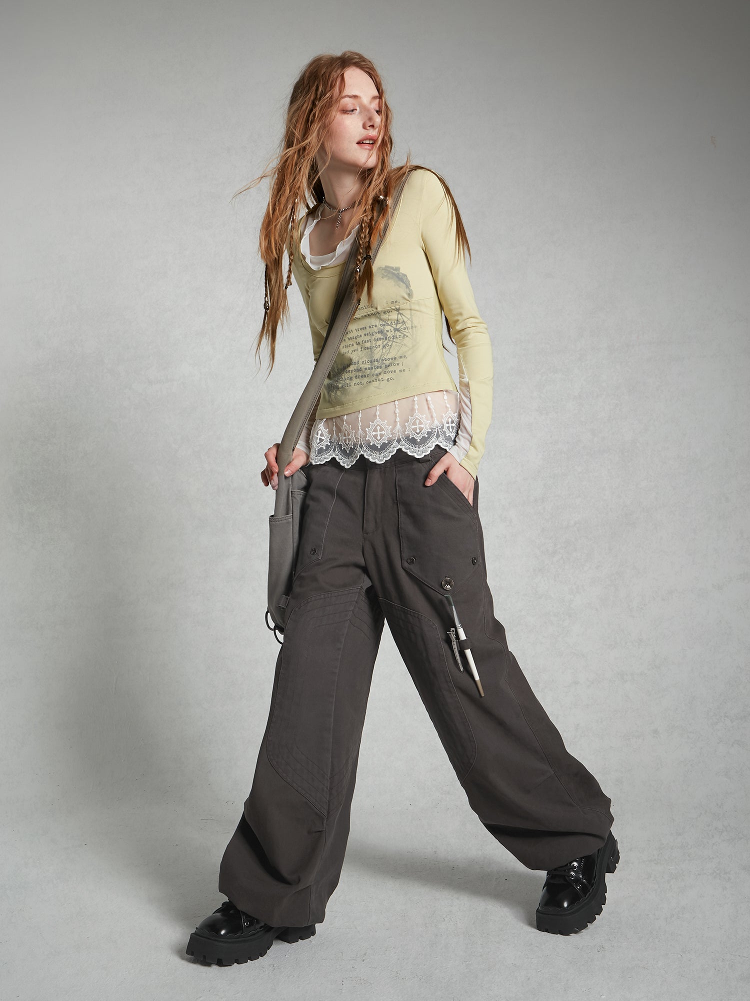 a full shot of a young woman modeling a casual outfit. she's wearing a pale yellow longsleeved top with lace trim, dark gray wideleg cargo pants, chunky black boots, and a gray shoulder bag. her long, reddishblonde hair is styled loosely. the background is a plain light gray. the overall style is somewhat grunge or alternative.