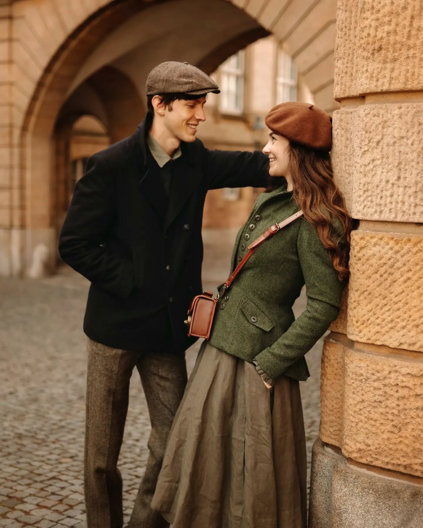 a romantic couple, dressed in vintage 1920s1930s style clothing, stands embracing near a stone archway. the man wears a newsboy cap and a dark coat, while the woman wears a beret and a green jacket with a long skirt. they are holding a small brown handbag. the setting is a cobblestone street with an old building in the background, creating a nostalgic and romantic atmosphere. the image evokes a sense of timelessness and classic style.