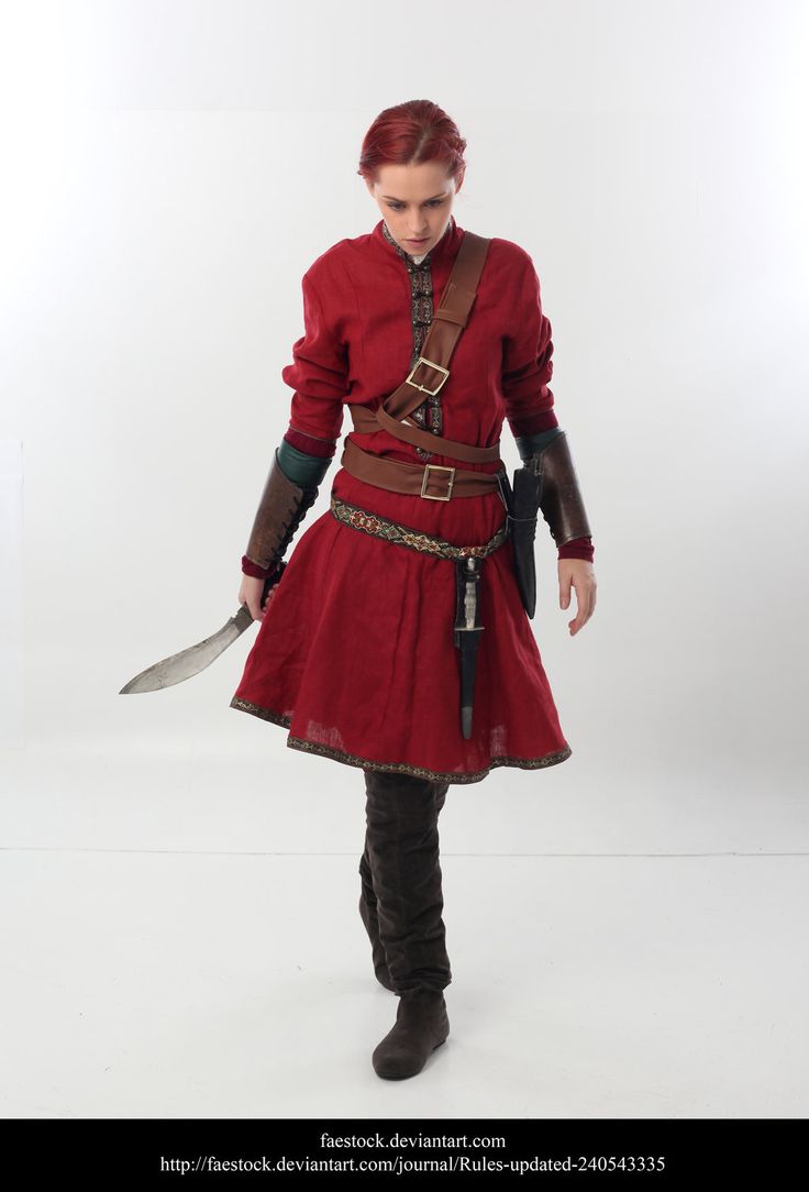 a woman in a red dress with leather accents and boots is standing in a white studio. she is holding a dagger in her right hand and a sword in her left hand. she is looking down at the ground.