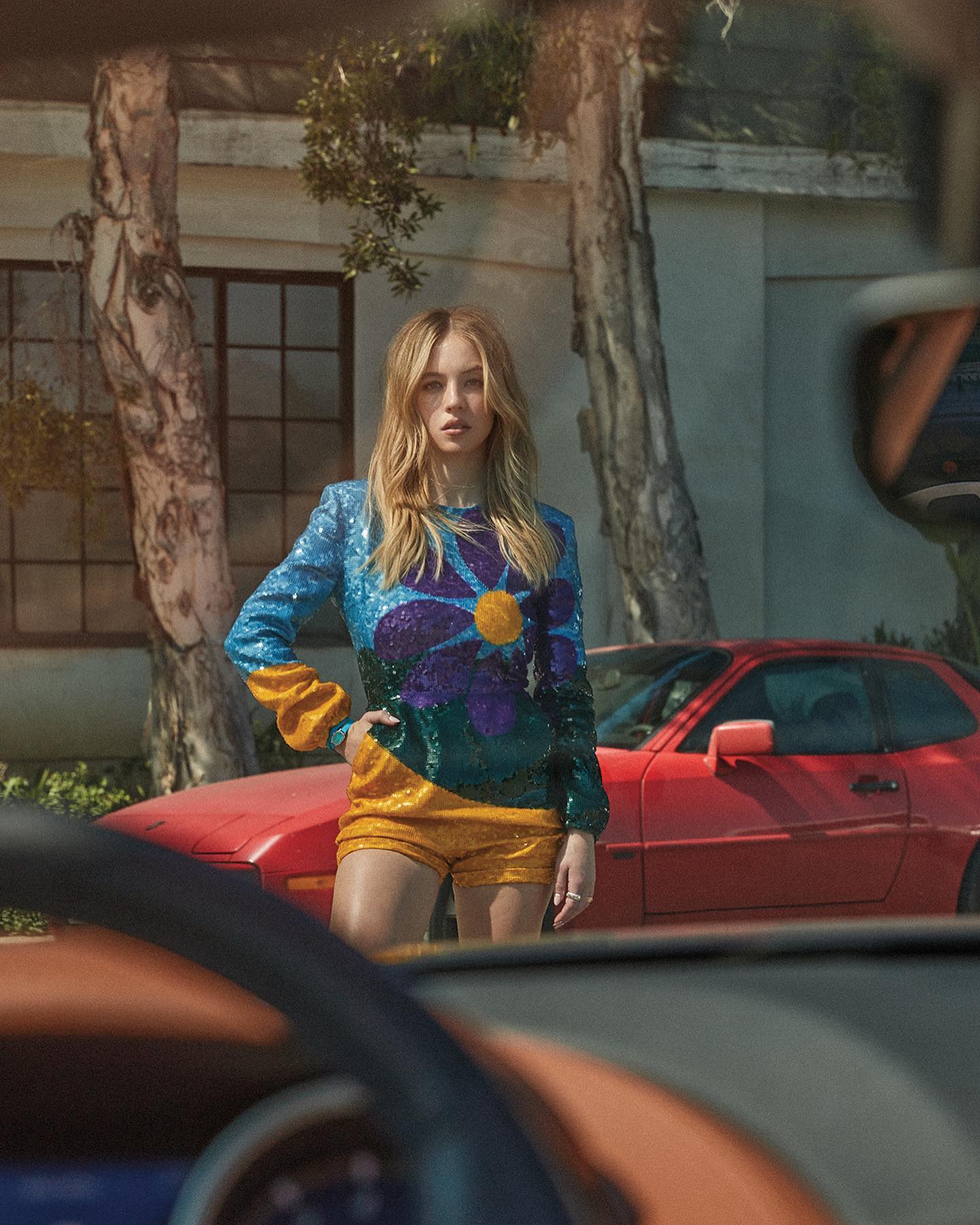 the image is a fashion photograph featuring a young blonde woman wearing a vibrant, sequined jumpsuit with a large flower design. the jumpsuit is primarily blue, green, and yellow. she is standing in front of a classic red porsche in what appears to be a sunny, residential californian neighborhood. the woman's pose is confident and stylish, and the overall aesthetic suggests a retro or vintage vibe. the image is taken from a slightly low angle, behind the wheel of another car which is partially visible, creating a framing effect. the background includes trees and a house, contributing to the scene's context. the image has a highquality, magazinestyle feel.