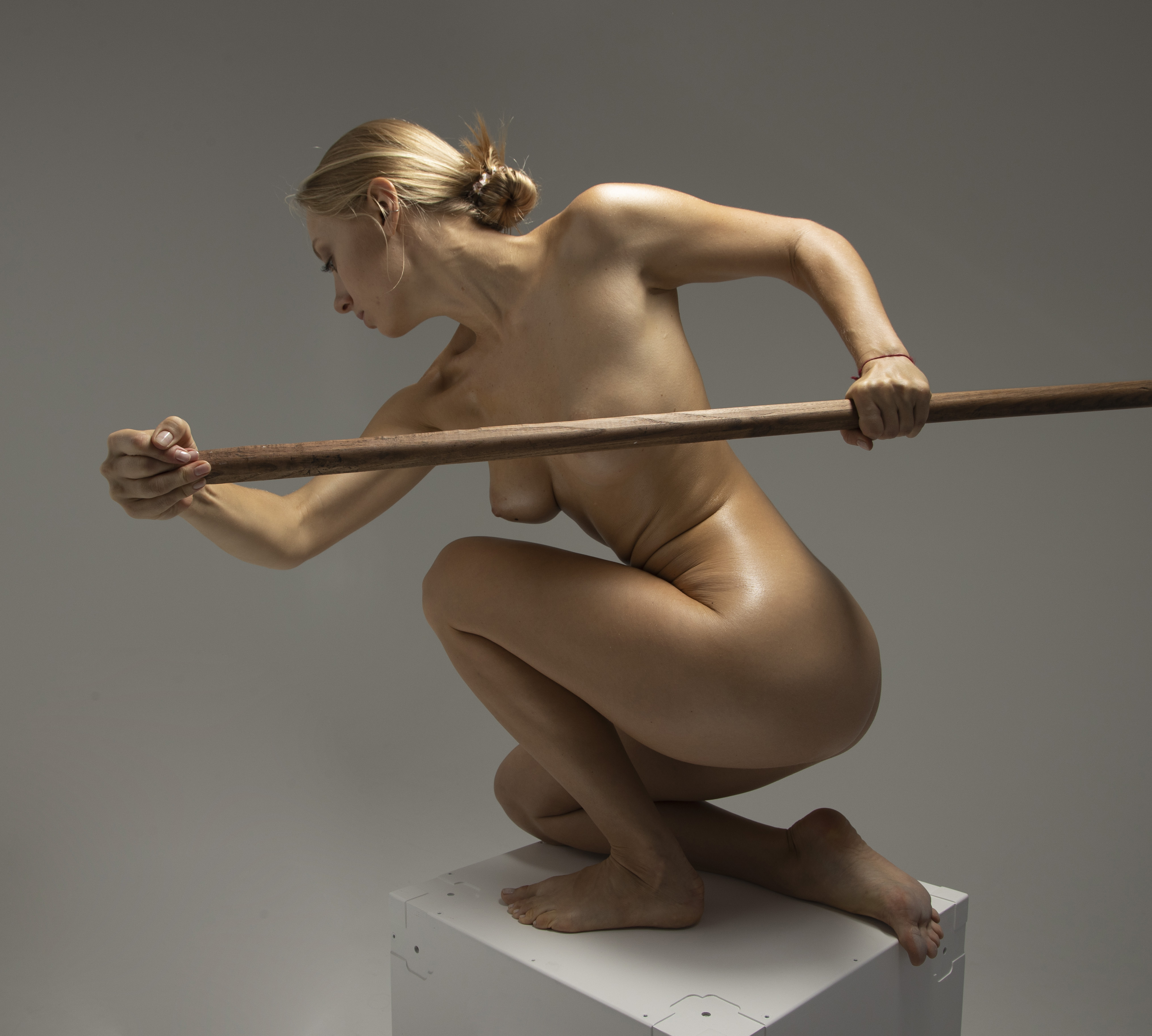 a nude blonde woman is squatting on a white cube in a studio setting, holding a wooden staff horizontally in front of her. she is posing with her head turned to the left and a determined expression. her body is illuminated, highlighting her musculature.