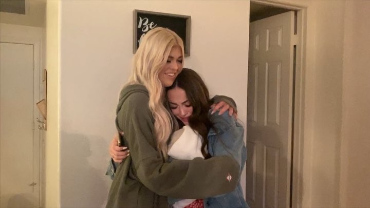 two women are hugging in an indoor setting. the woman on the left has long blonde hair and is wearing a green hoodie. the woman on the right has long brunette hair and is wearing a denim jacket. a white door is visible in the background, and there is a sign hanging on the wall above the women.