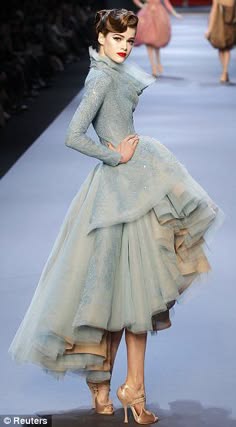 a fashion model walks the runway in a beautiful, light blue, multilayered dress with a ruffled skirt. the dress is made of tulle and features a fitted bodice. the model is wearing her hair up in a stylish bun and has a glamorous makeup look. she walks confidently in high heels,  showing off the elegant details of the dress. the background is a blurred runway with other models and audience members.  the overall atmosphere is chic and sophisticated.