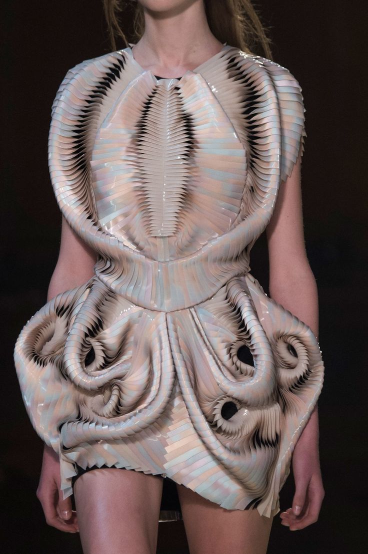 a model wearing a unique and avantgarde dress with a futuristic, abstract design. the dress is made of pleated fabric that creates a sculptural, organic effect. the dress is iridescent, shimmering with light and shadow. the model's pose and the overall style of the dress evoke a sense of elegance and beauty. this image highlights the creative and innovative nature of haute couture fashion.