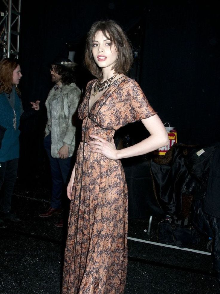 a young, elegant brunette model stands posing in a long, floral maxi dress backstage at a fashion show.  she is wearing a patterned dress and a necklace.  in the background, other people are visible, including two men, likely onlookers or other members of the fashion show's production. the overall atmosphere is dark and suggests a nighttime event. the model is the clear focal point of the image.