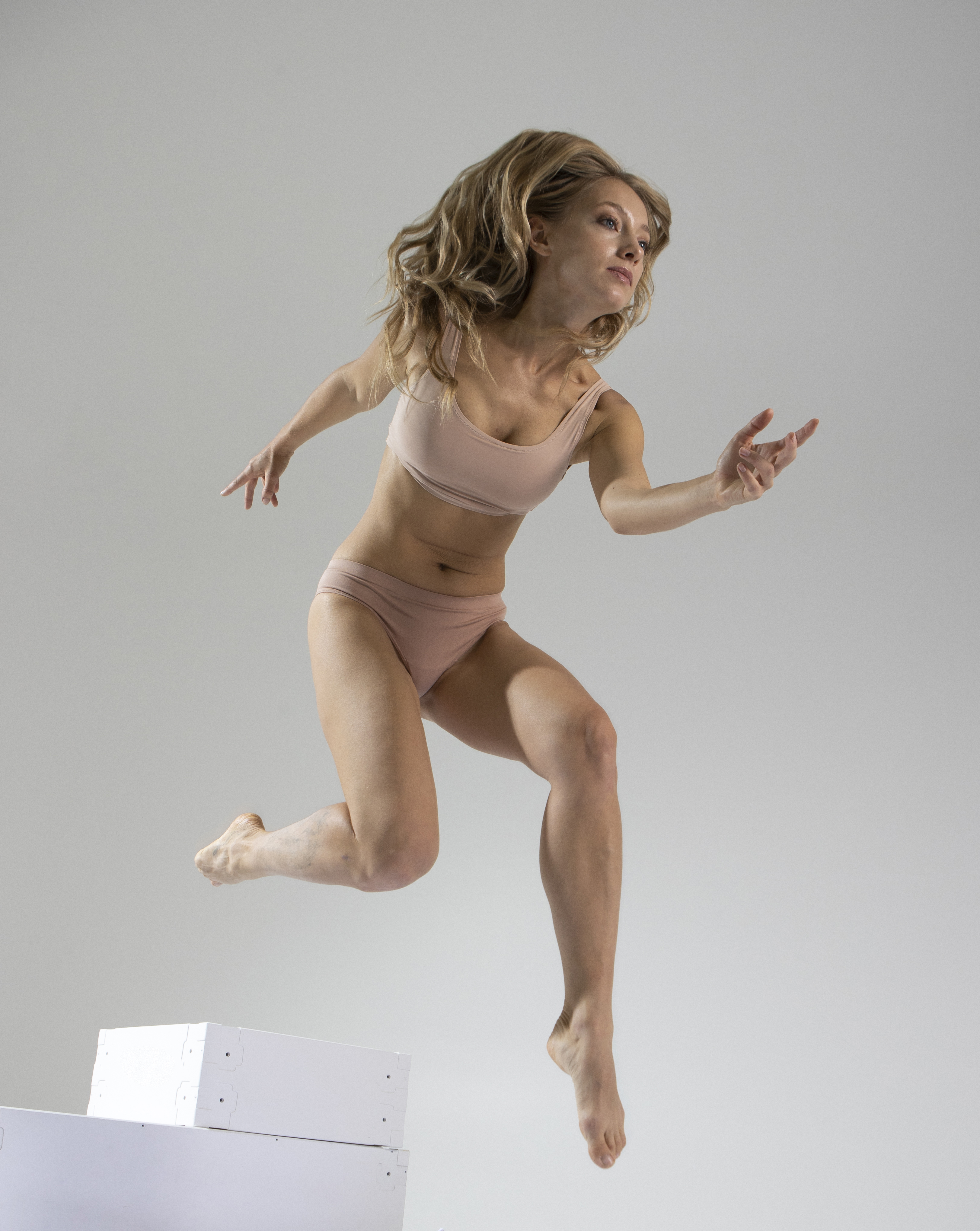 a woman with blonde hair is captured in a studio shot, jumping off of a stack of white boxes. she is wearing light pink lingerie and appears to be in motion, showcasing athleticism and fitness. the image conveys a sense of a healthy and dynamic lifestyle.
