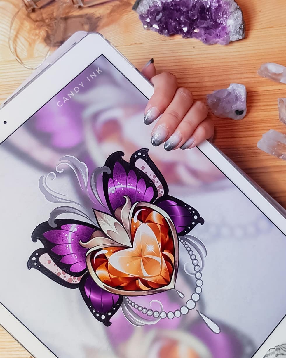 a drawing of a jeweled heart with butterfly wings, designed in a neotraditional tattoo style, is displayed on a tablet. the tablet is being held by a person with a colorful manicure.  purple crystals are scattered around the tablet on the table.