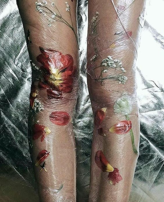 a closeup shot of a person's legs adorned with a temporary floral tattoo.  real flowers and petals are pressed against the skin and covered with a layer of clear plastic wrap, creating a translucent, ethereal effect. the flowers are mostly red and white, with some green stems and leaves visible.  the background is a crumpled silverygrey fabric. the overall aesthetic is artistic, delicate, and slightly unconventional.