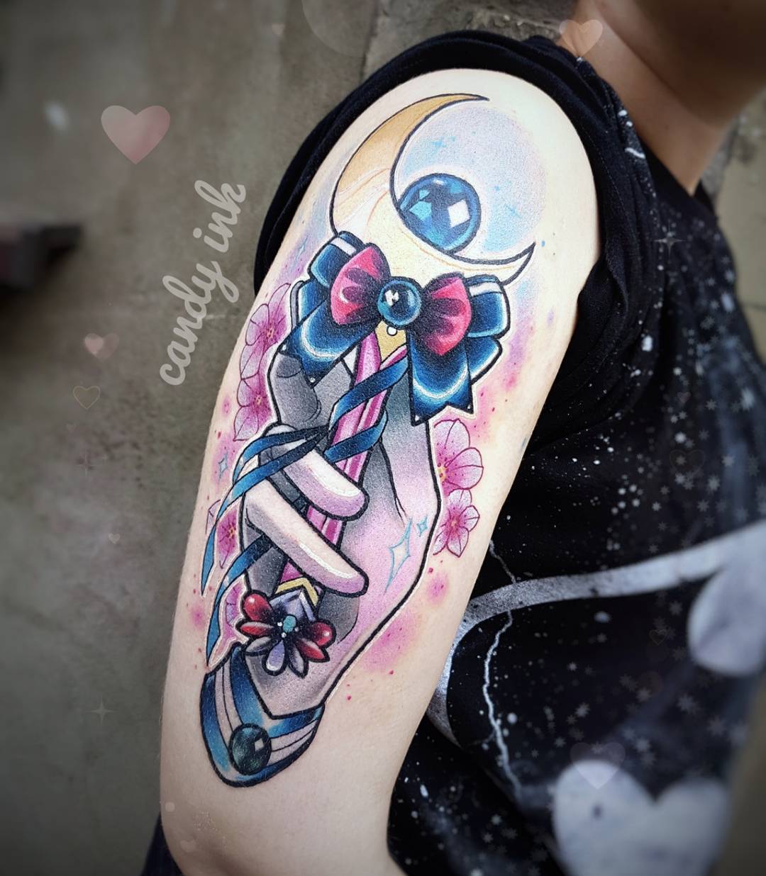 this is a tattoo on a person’s upper arm. the tattoo is of a hand holding sailor moon’s wand in front of a crescent moon. the moon and hand are done in a watercolor style with black outlines. there are also some cherry blossoms around the hand and wand. the hand is facing toward the viewer. the person who has the tattoo has light skin and freckles. they are wearing a black shirt with white polka dots. the background is blurry, but it looks like they are indoors. the image is lit with natural light.