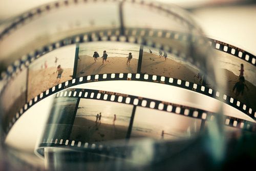 the image shows a closeup of a roll of analog film. the film has a series of photographs printed on it, showing people, mostly children, playing and running on a beach. the film is curved and overlapping, with the sprocket holes visible. the colors are muted and sepiatoned, giving the images a vintage feel.