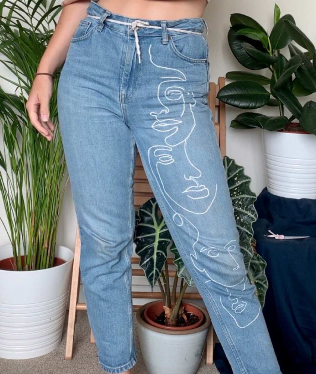 a person is modeling a pair of light wash denim jeans with a line art drawing of faces going down the leg. they are standing indoors in front of some houseplants. the photo is well lit with natural light.