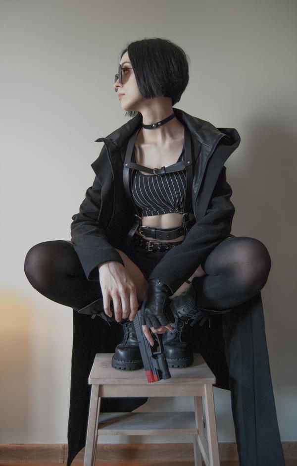 a young woman wearing a black leather jacket, choker, striped top, gloves, boots, and sunglasses is sitting on a small wooden stool with a gun in her hand. she has black hair and a serious expression on her face. the background is a plain white wall.