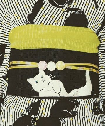 a closeup shot of a kimono featuring a black and white striped pattern with cat motifs. the obi (sash) is black with a large white cat design on it, accented by a yellow cord with three decorative beads. another yellow fabric is visible above the sash.