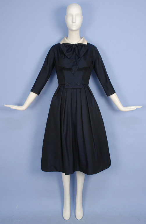 a navy blue vintage dress is shown on a white mannequin against a light blue backdrop. the dress, likely from the 1950s, features a fitted bodice, a full skirt, threequarter sleeves, and a prominent white collar with a bow. the soft lighting highlights the elegant drape of the fabric and the timeless silhouette of the garment.