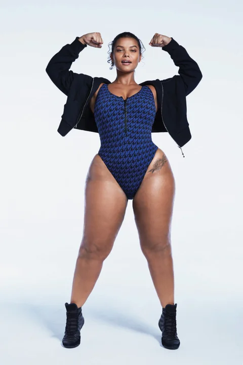 a plussize female model with toned physique confidently poses in a blue and black onepiece swimsuit with a front zipper. she wears a black zipup hoodie and black athletic shoes. her hair is pulled back, showcasing her strong features. the background is a bright white, typical of a studio photoshoot.