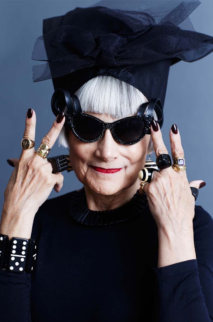 a closeup portrait of iris apfel, a renowned fashion icon, showcasing her signature style. she's wearing a black turbanstyle headwrap, large black sunglasses, and an array of bold statement jewelry, including multiple rings on each hand and a dominostyle bracelet.  her expression is confident and expressive, and her hands are playfully gesturing. the background is a simple, muted bluegrey, drawing focus to apfel's vibrant attire and personality.