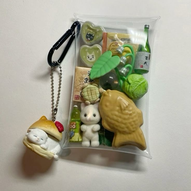 the image shows a charm/keyring hanging from a bag. the keyring has a cartoon cat in a bun. the bag is clear and contains a number of miniature items including heartshaped cat decorations, a melon treat, a lily of the valley flower, a japanese bottle, a head of cabbage, a turtle treat, a miniature bunny, and a fishshaped cake.