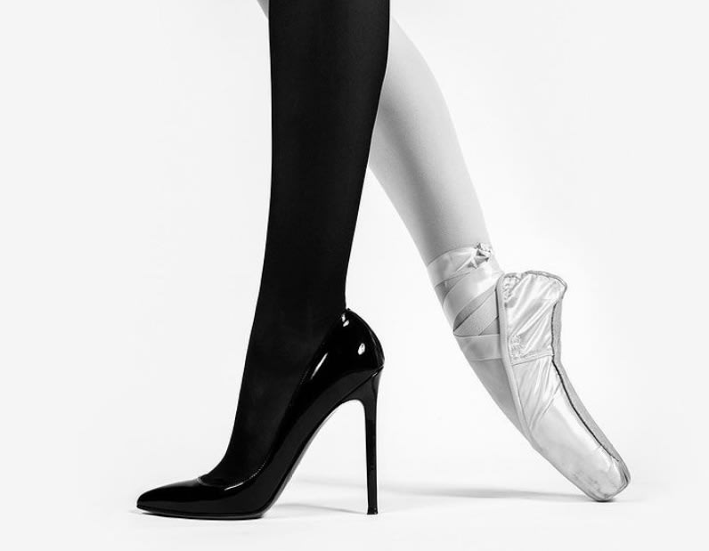 a woman's legs are shown wearing black pantyhose and high heels on the left, and white tights with a pointe shoe on the right. the image is black and white.