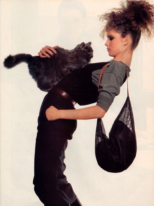 a woman, a model with styled hair is holding a black cat and standing with one arm raised. she is looking away. she is wearing a black dress and carrying a black bag.