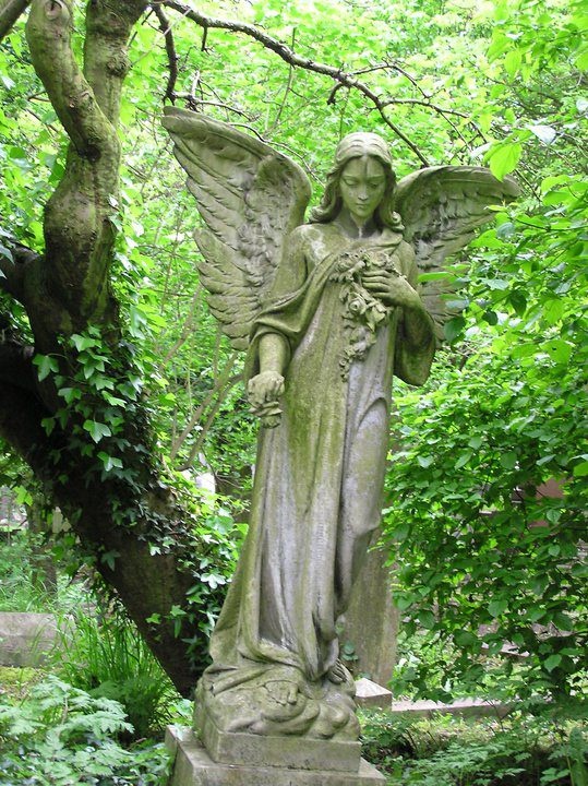 this image showcases a weathered, mosscovered angel statue standing gracefully amidst lush greenery. the angel, with delicate wings unfurled, holds a bouquet of flowers close to its chest, its head bowed slightly in a gesture of mourning or contemplation. the statue is positioned in a cemetery, with leafy trees and overgrown foliage forming a serene and tranquil backdrop. the soft, natural lighting casts gentle shadows, enhancing the atmosphere of peace and remembrance.