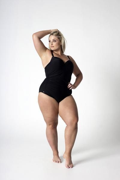 a woman in a black swimsuit stands with her weight on her right leg and hand on her hip, posing in a studio against a white backdrop. she is looking off to the side and has a confident expression.