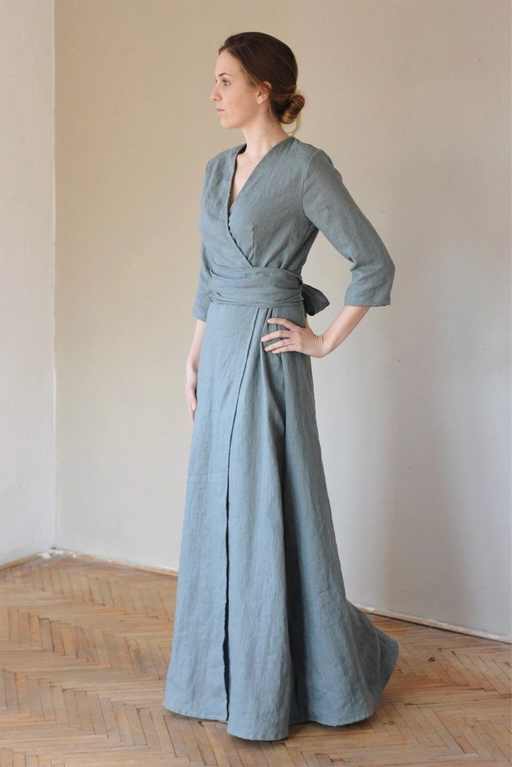 a woman is standing sideways in a long blue wrap dress. the dress is made of linen and has threequarter sleeves. the dress falls to the floor and has a slit on one side. the woman has her hair pulled back in a bun and is looking to the left. the background is a white wall and a wooden floor. the lighting is soft and natural.