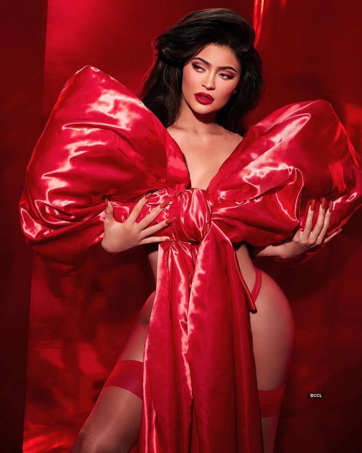 the image showcases kylie jenner in a striking photoshoot. she's dressed in a voluminous, red satin bowshaped garment that resembles a large gift or present.  the fabric is shiny and draped dramatically around her body, which is partially visible beneath the translucent garment. kylie is wearing red lipstick, matching her attire,  and has her dark hair styled in soft waves. the background is a deep crimson, complementing the overall red theme. her pose is sensual and confident. the overall aesthetic is glamorous and suggestive, possibly themed for valentine's day or a similar holiday.