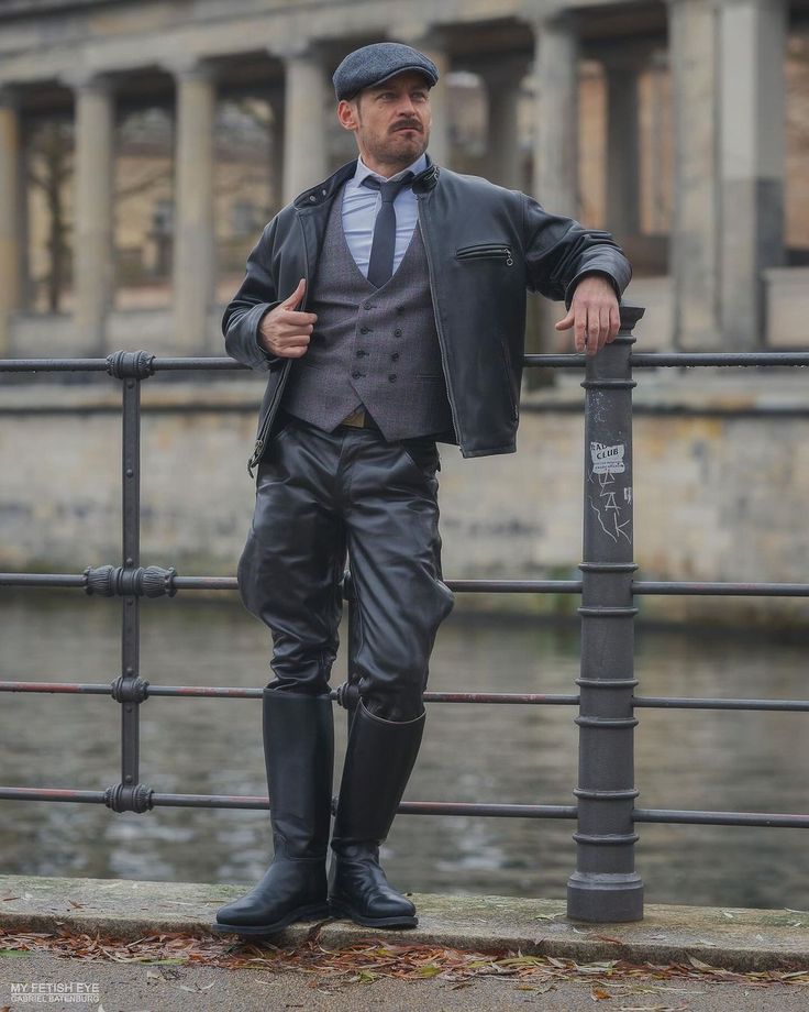 the image showcases a stylishly dressed male model leaning against a metal railing on a bridge. he is wearing a black leather jacket,  black leather pants, and tall black riding boots. underneath, he sports a grey waistcoat and vest, a light blue shirt, and a dark tie  a threepiece suit ensemble. he's also wearing a dark newsboy cap.  the background is a blurry but noticeable city scene with classical architecture and water visible beyond the railing. the overall style evokes a vintage, possibly 1920s1930s, or gangsteresque aesthetic. the model appears confident and stylishly posed. the image has a moody, somewhat dramatic lighting.