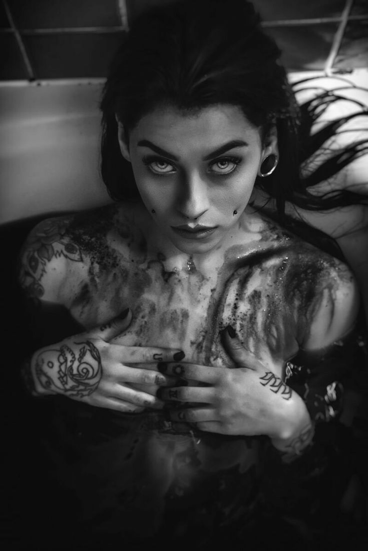 a black and white portrait of a young woman in a bathtub. she has long, dark hair and is wearing tattoos. her