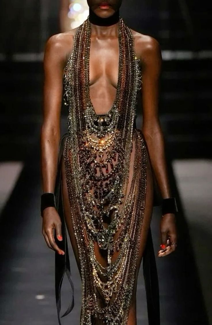 a darkskinned model is walking on a runway wearing a beaded dress and multiple beaded necklaces. she is wearing a black choker and thick black cuffs on both wrists. the dress is almost entirely beads, with nude undergarments visible underneath. black ribbons are attached to the sides of the dress at the waist. the model has dark hair and red nail polish.