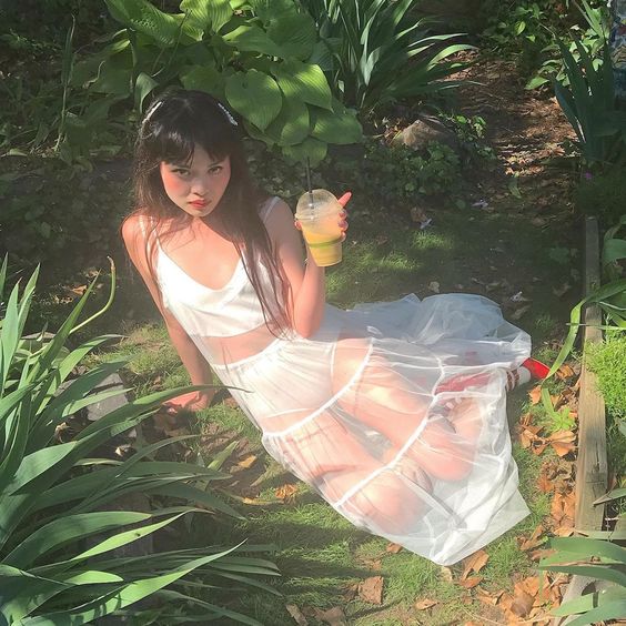 a young woman with long black hair is sitting in a lush green garden, wearing a white sheer dress. she is holding a beverage cup in one hand and is looking directly at the camera. the photo is taken from a high angle, showcasing the woman and the surrounding greenery. the lighting is natural and sunny, creating a bright and airy feel.