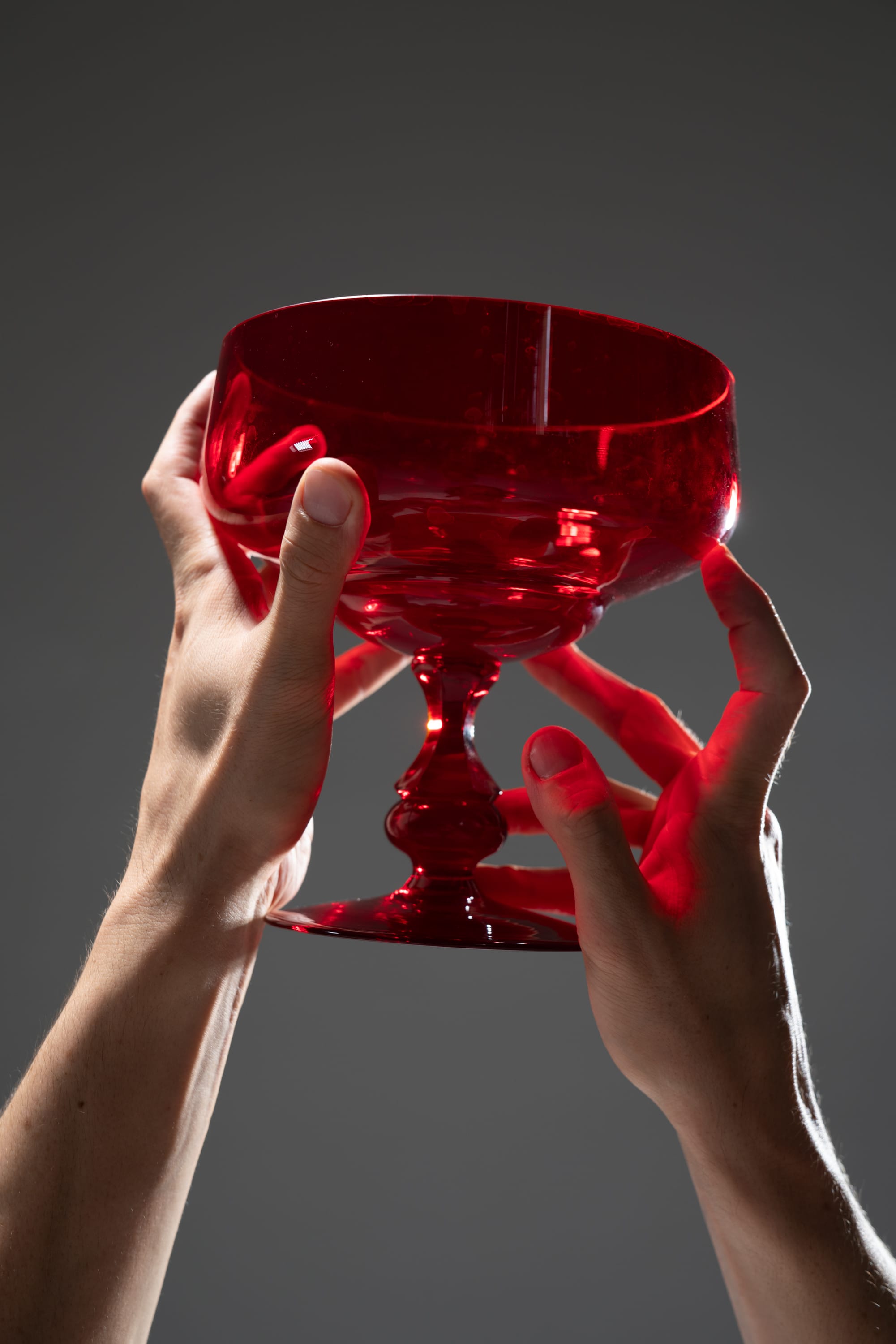 a pair of hands delicately holds a vibrant red glass goblet against a muted gray backdrop, the studio lighting casting dramatic shadows and highlighting the translucent beauty of the glassware.