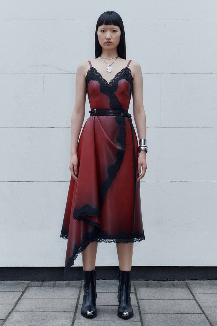 a slender woman poses in front of a textured white wall, dressed in a striking red leather dress with black lace accents. the dress features a thin black belt that cinches her waist. on her feet, she wears sleek black boots with silver detailing. she accessorizes with a silver necklace and a wide silver bracelet. her black hair is styled with blunt bangs.