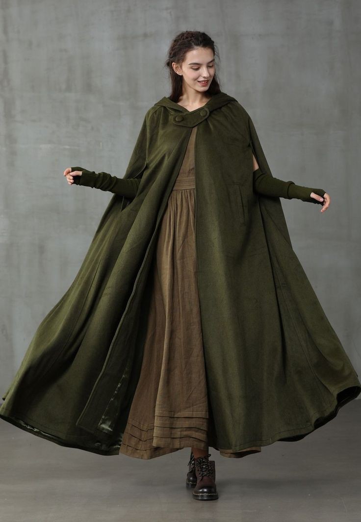 a woman stands in a fulllength shot, wearing an olive green medievalstyle cape with a hood and thumb hole sleeves. underneath, she has on a long brown skirt and dark brown boots. the cape has buttons at the neck and drapes down to the floor. the background is a plain gray.