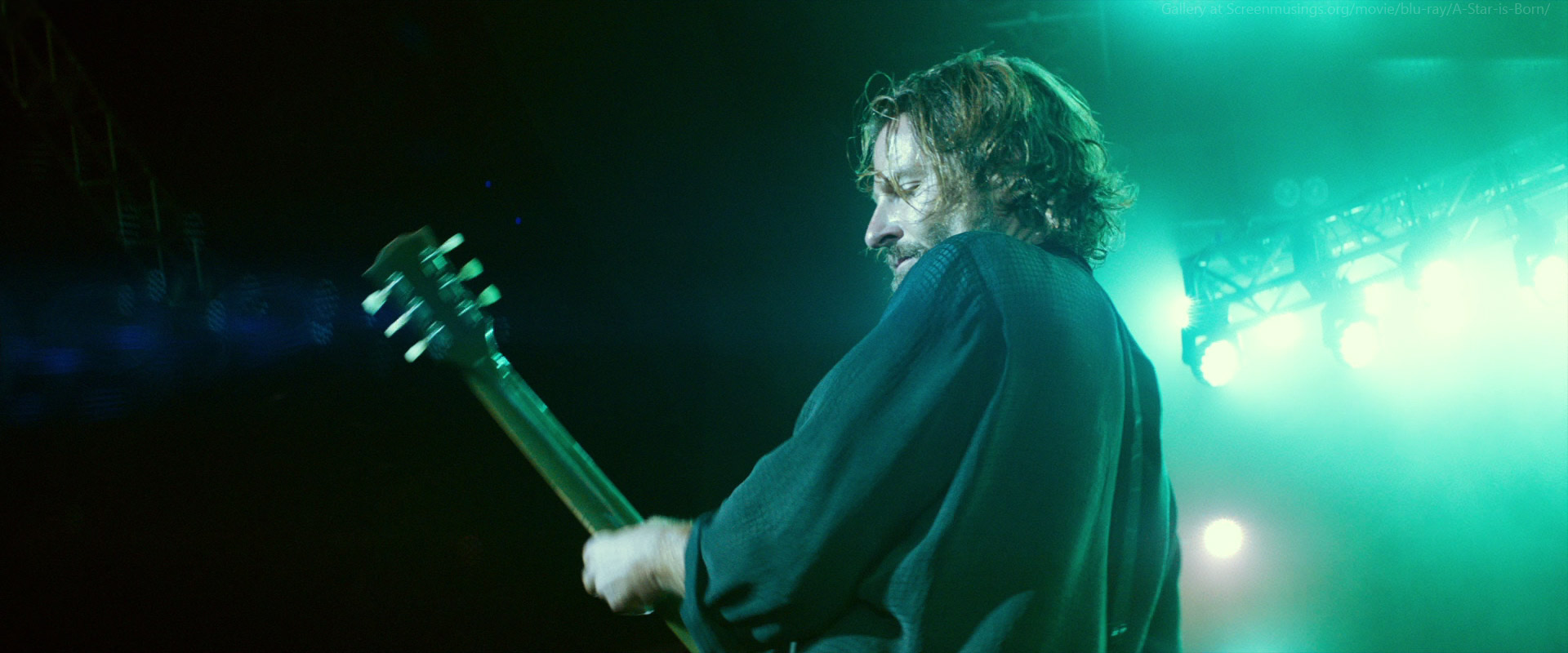 a man with long brown hair plays an electric guitar during a rock and roll concert. the man is wearing a dark blue shirt and is lit from behind by a spotlight. the stage is dark and smoky and filled with more lights.