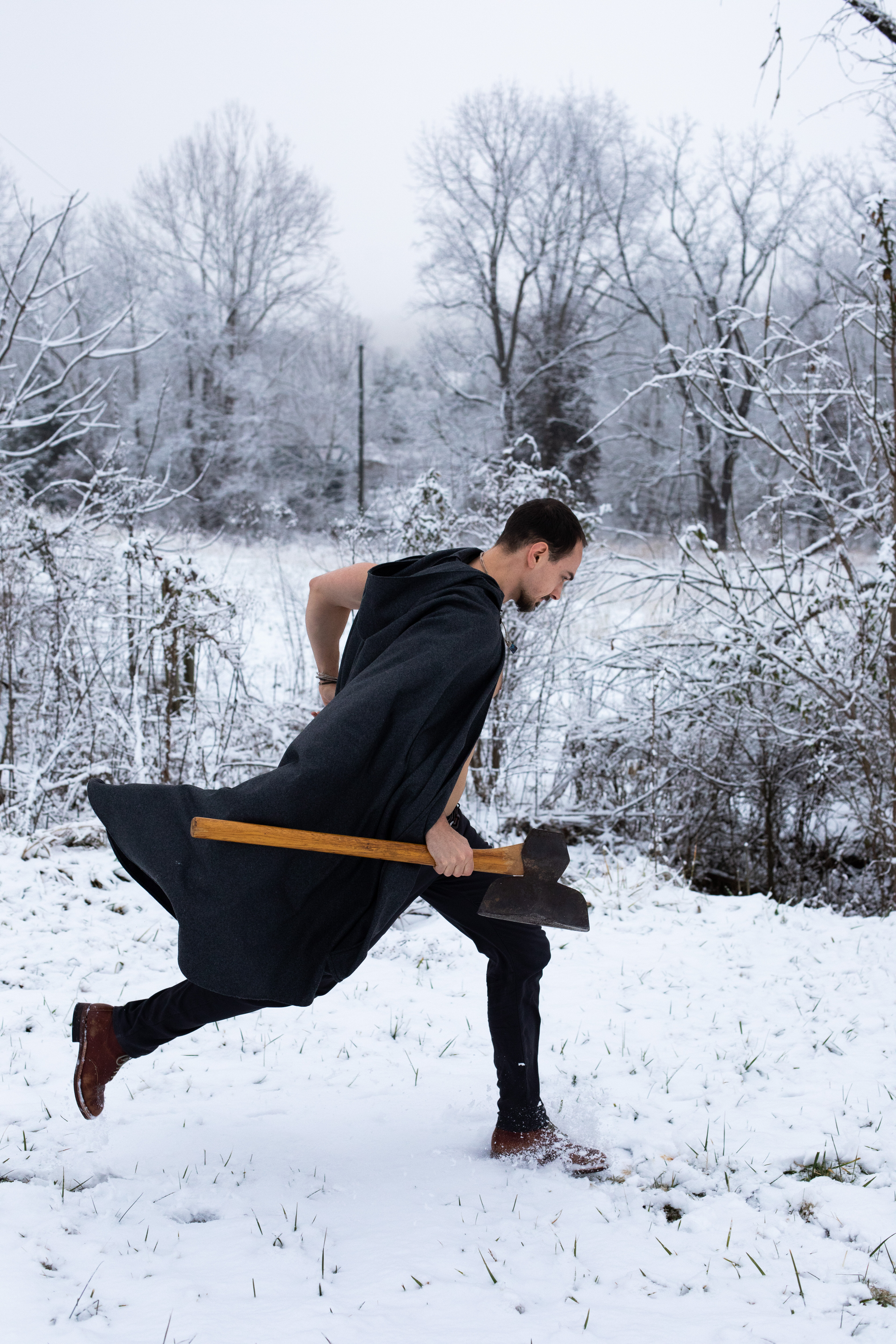 a young man, clad in black pants and a flowing black cape, sprints through the snow. he is barechested despite the wintery setting. in his hands, he carries a large, doubleheaded axe, held firmly as he charges forward. the snowy forest provides a stark, monochromatic backdrop, with bare trees reaching towards the overcast sky. his intense focus and the dynamic pose suggest action and purpose.