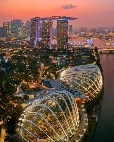 the image is a stunning aerial shot of singapore's iconic cityscape at sunset or twilight.  the main focus is on the marina bay sands hotel with its distinctive threetower design and rooftop infinity pool, prominently displayed against the backdrop of a vibrant, colorful sky.  in the foreground, the breathtaking gardens by the bay, specifically the flower dome and cloud forest conservatories, are showcased with their unique biodome structures beautifully illuminated at night. the image shows the juxtaposition of modern architecture and lush greenery, with the city lights reflecting on the water surrounding marina bay. the overall effect is a captivating view of singapore's unique blend of natural beauty and urban development.