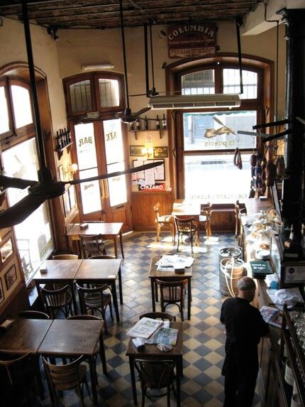 the image is an overhead shot of a traditional, dimly lit bar or cafe interior. the space features vintage wooden furniture, including numerous small tables and chairs arranged throughout the room. the floor is a classic checkered tile pattern.  large windows let in some natural light, showcasing the building's age and character.  the ceiling has multiple ceiling fans. there's a bar area in the back with hanging cured meats and bottles visible on shelves. a sign reading "columbia" is visible on the wall. a bartender is seen tending to a customer. the overall atmosphere is rustic, traditional, and inviting, suggesting a spanish or european setting.