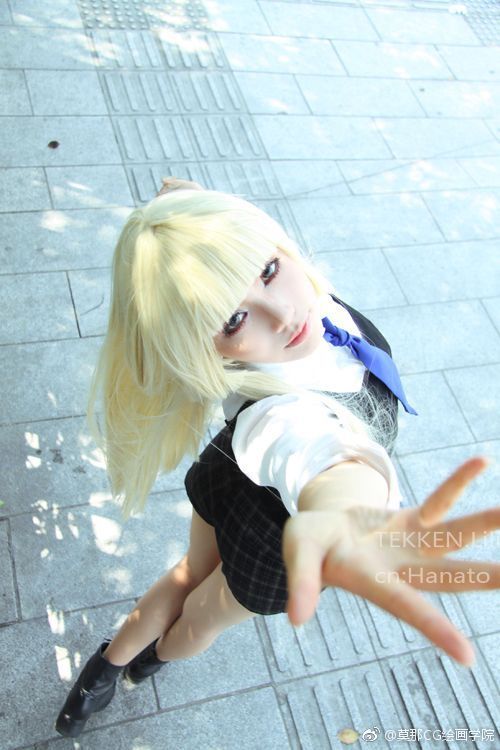 a blonde haired girl in a school girl cosplay with a black plaid skirt, white shirt, and blue tie looks at the camera with a cute smile. she is posed with one hand outstretched toward the viewer. the backdrop is of an outdoor street scene with grey concrete and light grey bricks on the sidewalk.