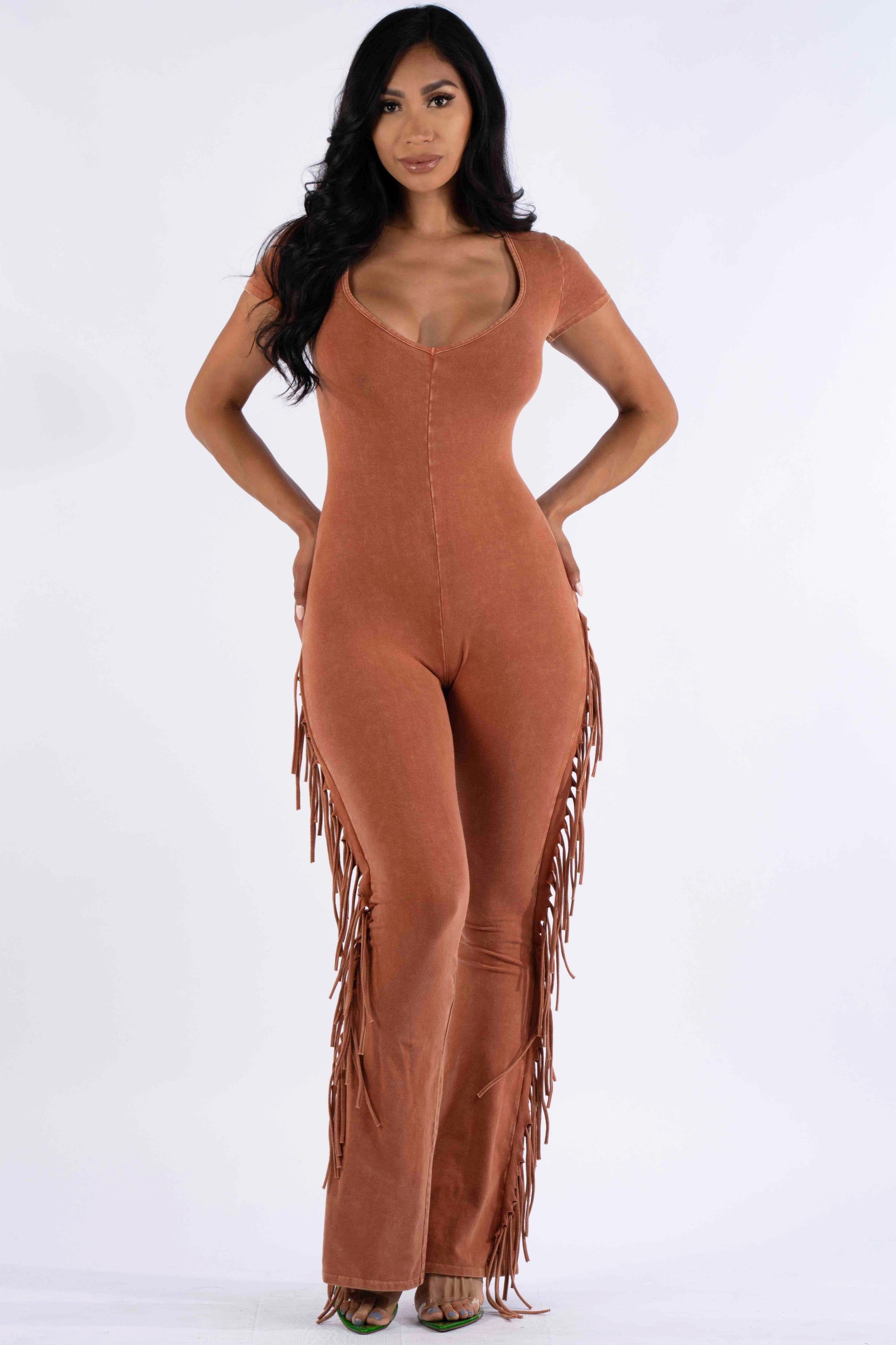 a fulllength studio shot of a female model showcasing a terracottacolored jumpsuit. the jumpsuit features short sleeves, a vneckline, a fitted silhouette, and fringe detailing along the sides of the legs. the model is posing with her hands on her hips, showcasing the garment's style and fit. the background is a plain white, focusing attention on the model and her attire.  the overall style of the jumpsuit is casual yet stylish.