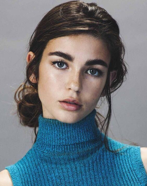 loseup portrait of a young woman with brown hair styled in a low bun. she's wearing a teal turtleneck sweater and has minimal makeup, showcasing her natural beauty.  her expression is serious and her gaze is directed towards the camera. the background is a simple, neutral gray.
