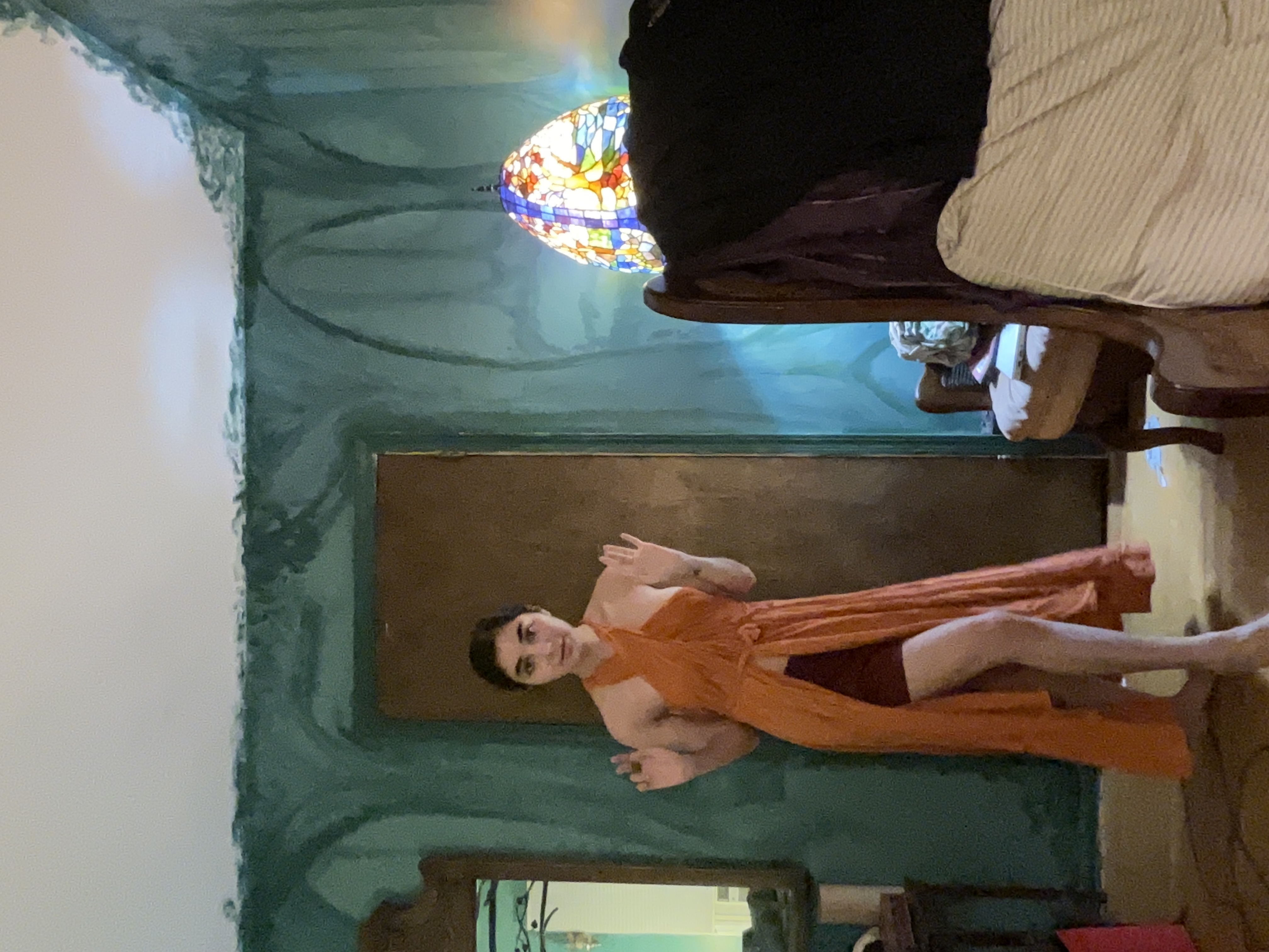 a young woman is standing in a room with teal walls and antique furnishings. she is wearing a flowing orange wrap dress that is highlow in style.  she's posing casually with her hands slightly raised.  in the background, there is a vintagestyle chair and a stainedglass lamp. the overall aesthetic is vintage or bohemian. the room appears to be a bedroom.