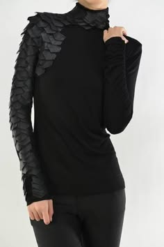 a woman wearing a black longsleeved turtleneck with leather scale details on the shoulders, posed with her right arm bent at the elbow. her hair is obscured in the image. she is wearing black pants and a black shirt. the background is a light solid color.