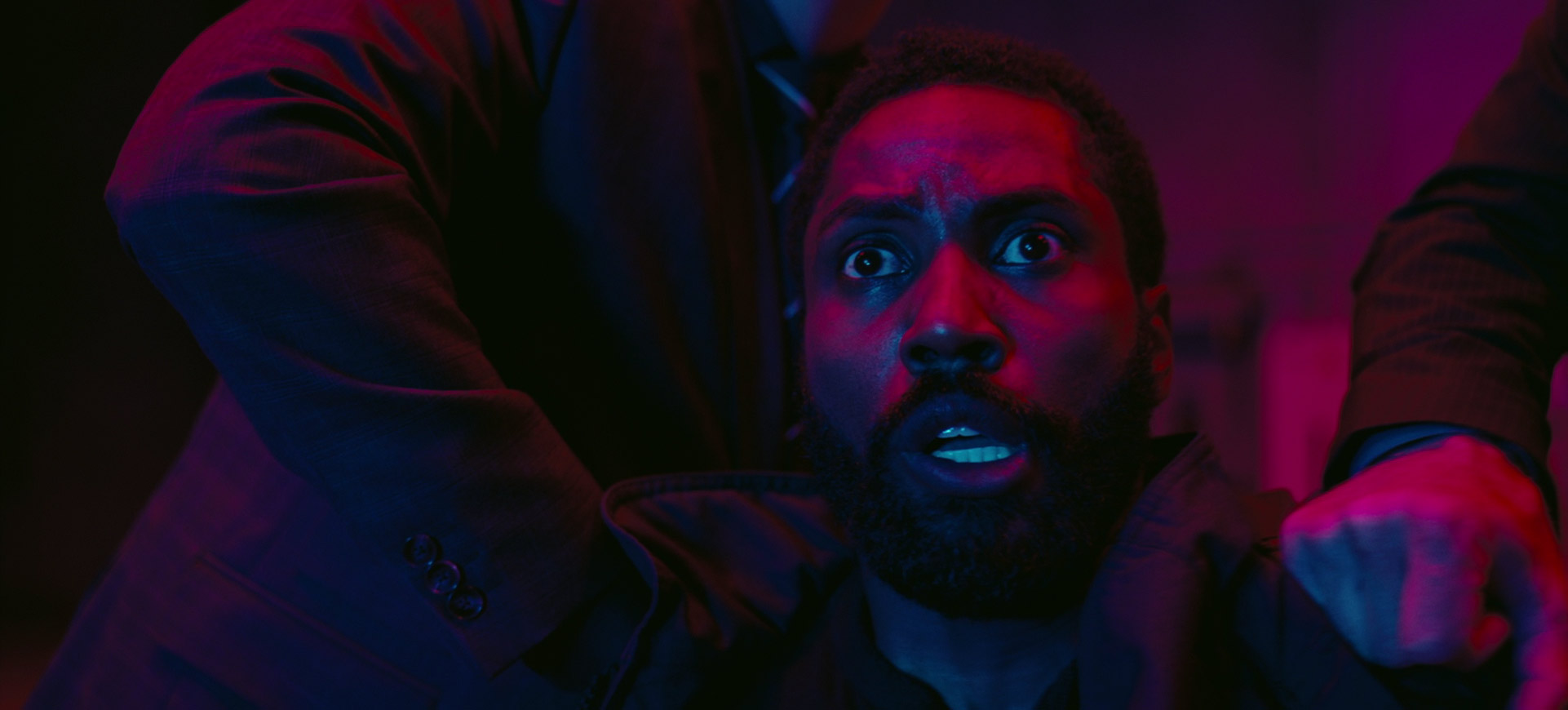 a closeup image of a man in a suit with a scared expression on his face. he is illuminated with blue and red light and a hand is gripping his jacket in a tense manner.