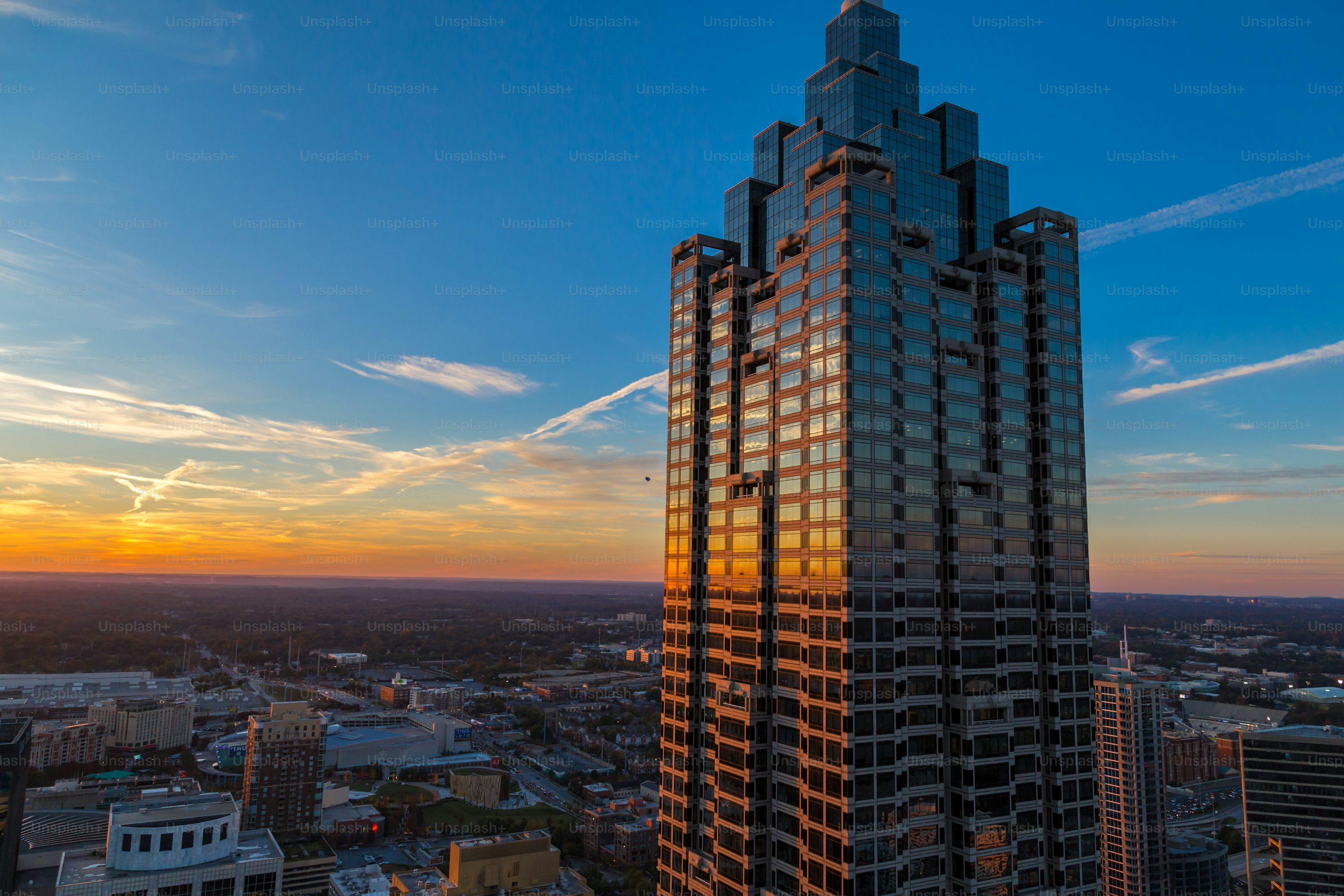 the image showcases a tall, modern skyscraper dominating the skyline of a city at sunset. the building, primarily made of glass, reflects the warm hues of the setting sun. the sky is a mix of blue and orange, with streaks of clouds adding to the scenic view. the cityscape is visible below, with other buildings and streets visible in the distance. the overall composition captures the beauty of urban architecture against the backdrop of a vibrant sunset.