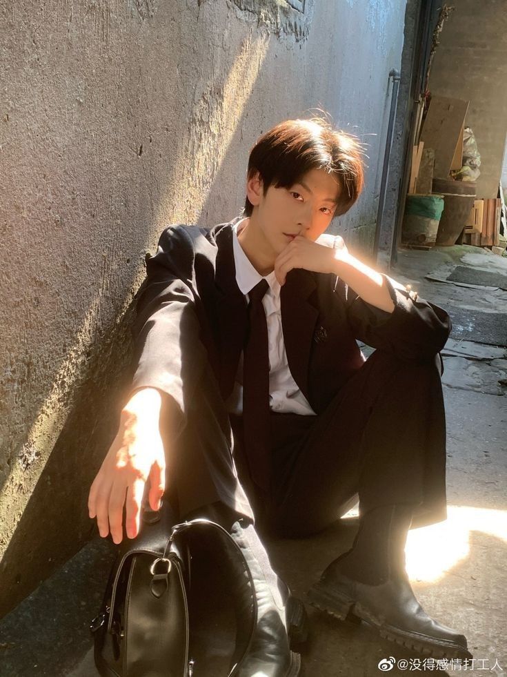 a young man in a black suit and tie poses in a sundrenched alleyway. he is sitting on the ground, leaning against a wall, with one hand on his knee and the other reaching towards the camera. he has a black leather bag next to him. the image is welllit, with natural sunlight creating strong shadows and highlights.