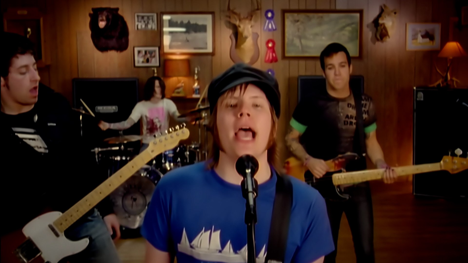 a fourperson band is playing in a woodpaneled room. the lead singer, wearing a blue tshirt and a black cap, is singing into a microphone. the guitarist on the left is wearing a black tshirt, and the guitarist on the right is wearing a green tshirt. the drummer is in the background. there are various decorations on the walls, including a mounted bear head, a deer head, and framed pictures. the lighting is bright and artificial. the image is taken from eye level.