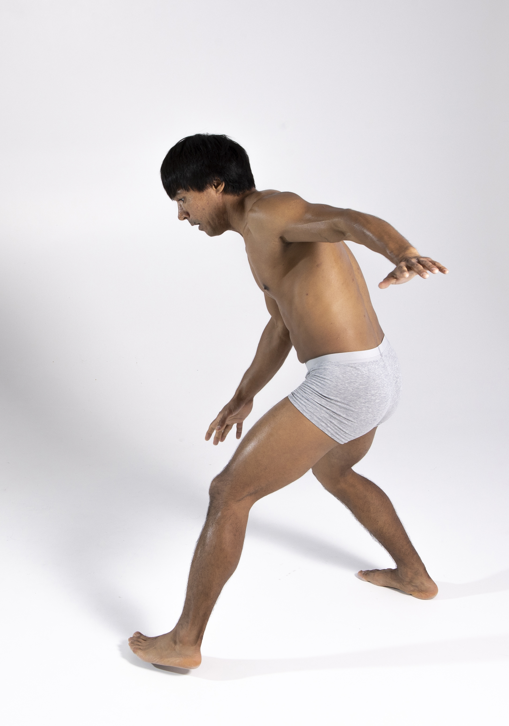 a full shot of a barechested man wearing gray underwear standing on a white background, looking down with his arms slightly raised in a gesture of balance or anticipation. his body is forwardleaning and he is barefoot, with one leg slightly forward than the other, in a studio setting under bright lighting.