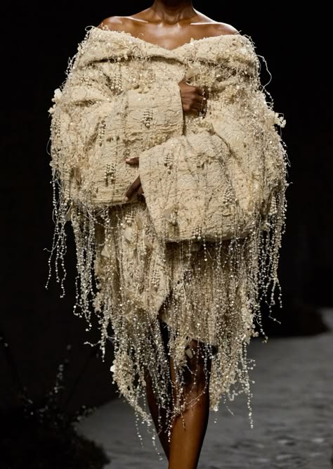a model walks the runway in a white beaded dress with fringe detailing. the dress is off the shoulder and features a high neckline. the model's hair is styled in a simple updo, and her makeup is minimal. the dress is very ornate and intricate, with a lot of detail in the beading and fringe. the model is wearing a lot of jewelry. the background is dark and out of focus.