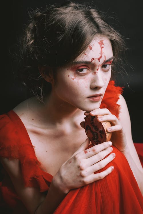 closeup portrait of a young woman with dark hair styled in an updo. she is wearing a red dress and her face and hands are stained with what appears to be blood. she holds a pomegranate in her hands. the background is dark and the overall mood is dramatic and mysterious, suggesting themes of tragedy, grief, or sacrifice. the image has a vintage or antique feel. the makeup is emphasized, enhancing the dramatic effect.  the woman's expression is one of sadness or contemplation.
